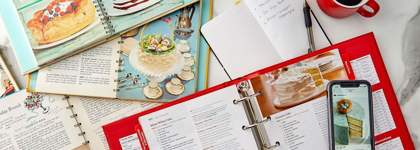 Multiple Betty Crocker Cookbooks, a white notepad, cup of coffee and a cell phone displaying a image from social media