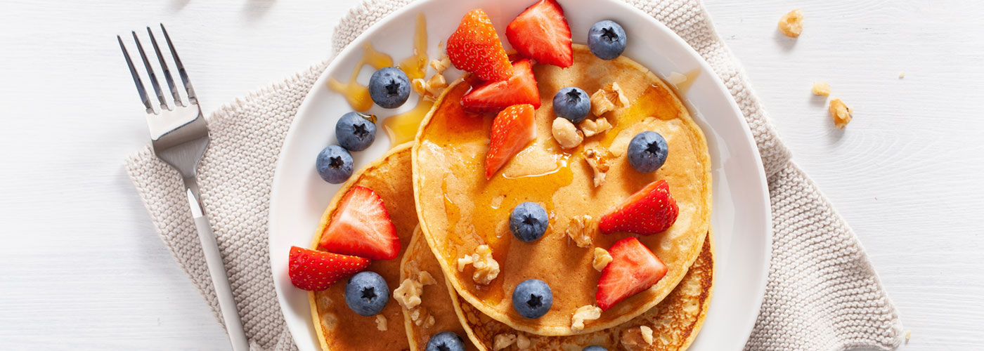 Lifestyle image of a plate of Pancakes topped with nuts, strawberries, blueberries and syrup. On a white plate with a silver fork.