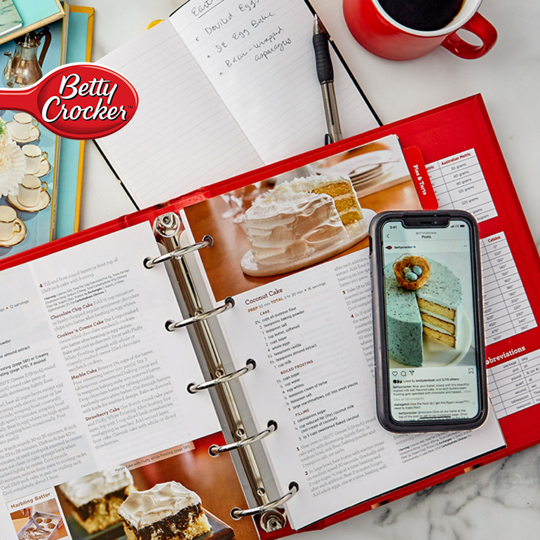 Lifestyle image of multiple Betty Crocker Cookbooks, a white notepad, cup of coffee and a cell phone displaying a image from social media