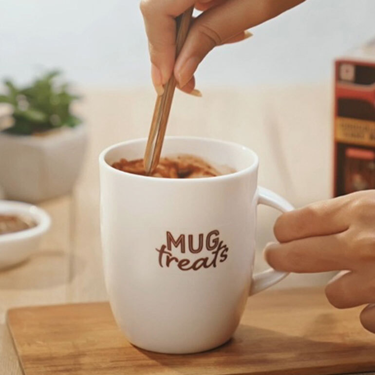 A white mug with text on it saying "Mug Treats" and a hand mixing the content inside