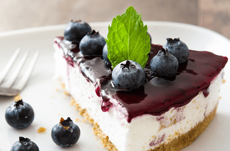 A slice of Cheesecake on a white plate with blueberries and a mint leaf on top