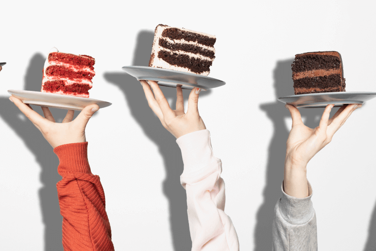 Three slices of cake being held up by three hands. One is chocolate with vanilla frosting, one is chocolate with chocolate frosting and one is strawberry with vanilla frosting