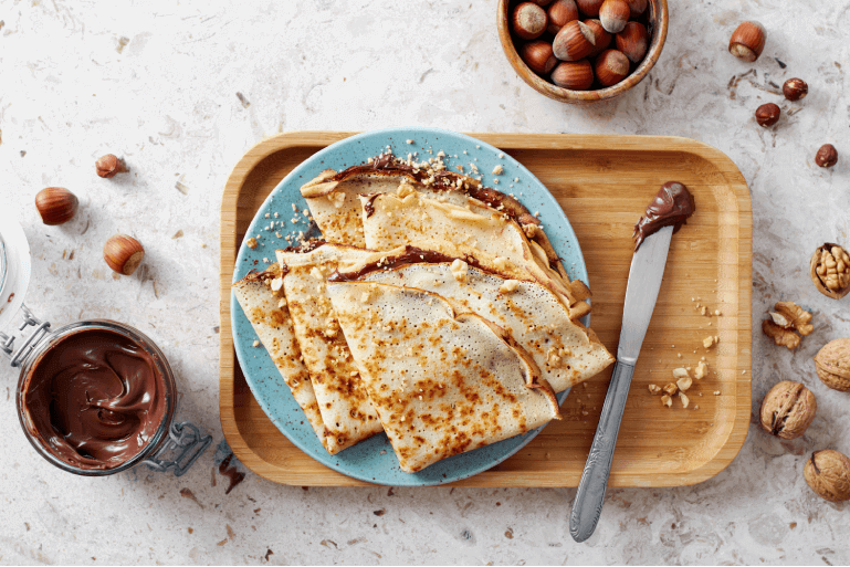 A lifestyle image of Chocolate Spread & Banana Crepes on a blue plate with a knife with chocolate sauce on it