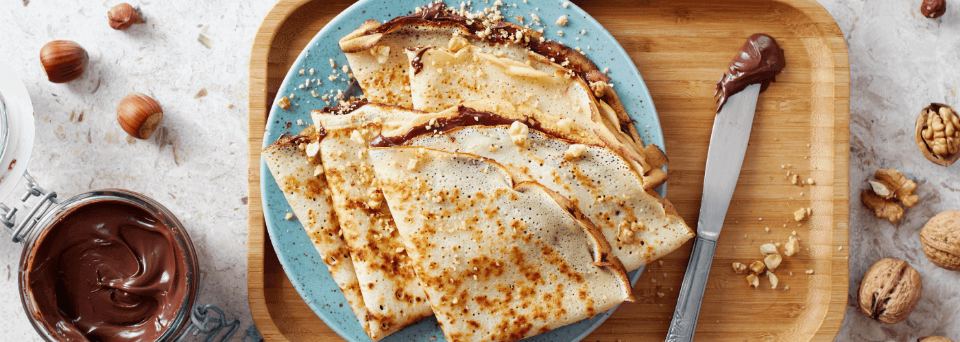 A lifestyle image of Chocolate Spread & Banana Crepes on a blue plate with a knife with chocolate sauce on it