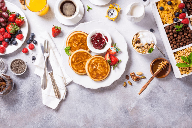 A table of various food such as pancakes, fruit, honey and nuts on white plates and silver silverware