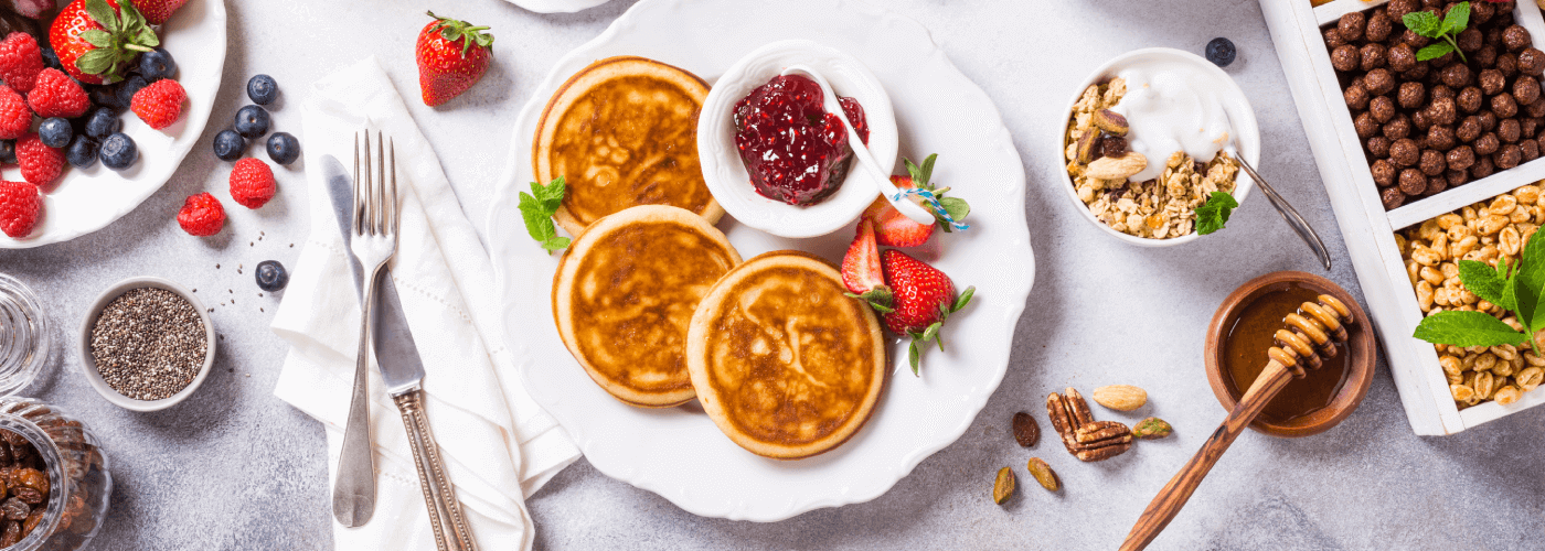 A table of various food such as pancakes, fruit, honey and nuts on white plates and silver silverware